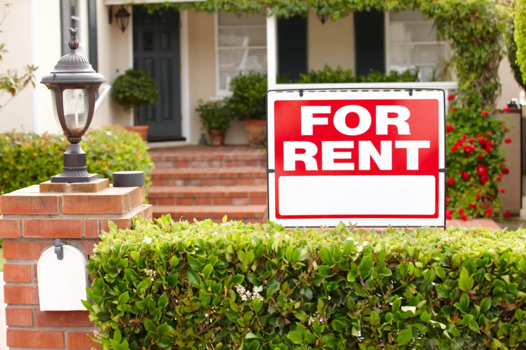 Red For Rent sign in front of a single-family home with a brick walkway and hedges, marking the case for renting out your house instead of selling